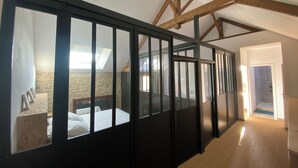 Una habitación con techo inclinado, vigas de madera a la vista, una cama con ropa blanca y un ventanal con marcos negros.