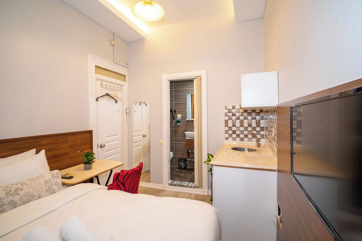 A compact hotel room with a bed, bedside table, and a small kitchenette.
