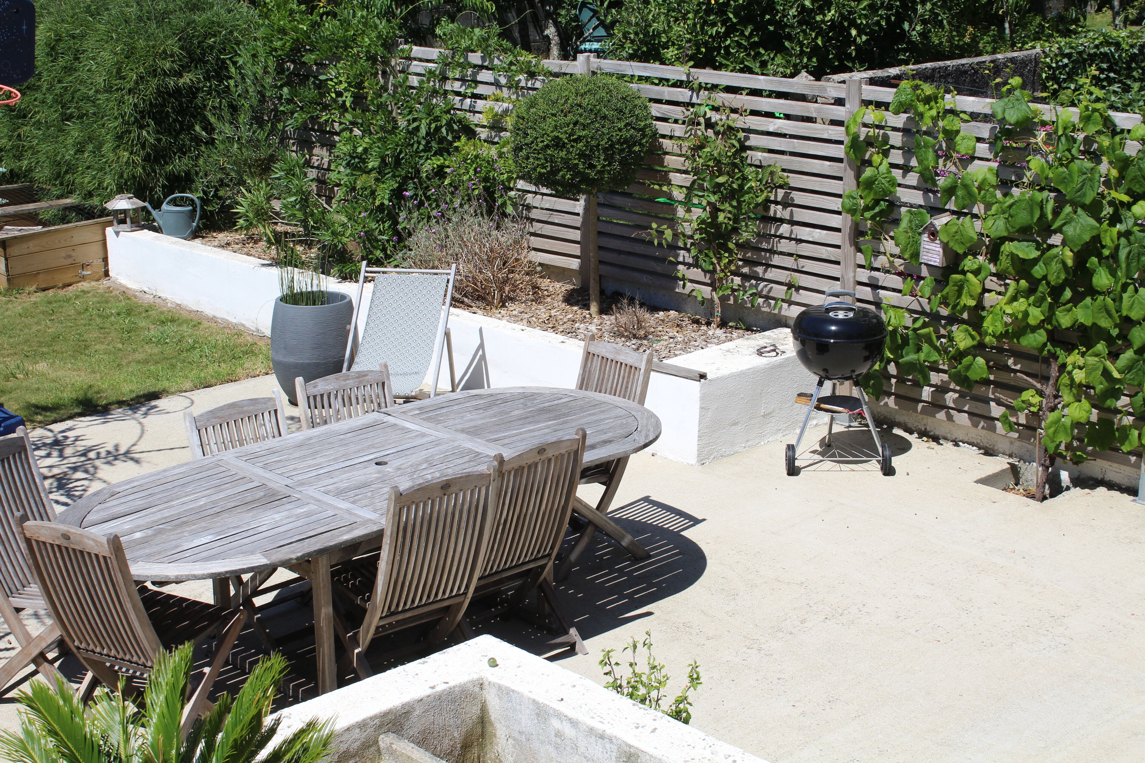 Juego de mesa y sillas de madera al aire libre, con una parrilla de barbacoa y un jardín con plantas y cerca de madera.