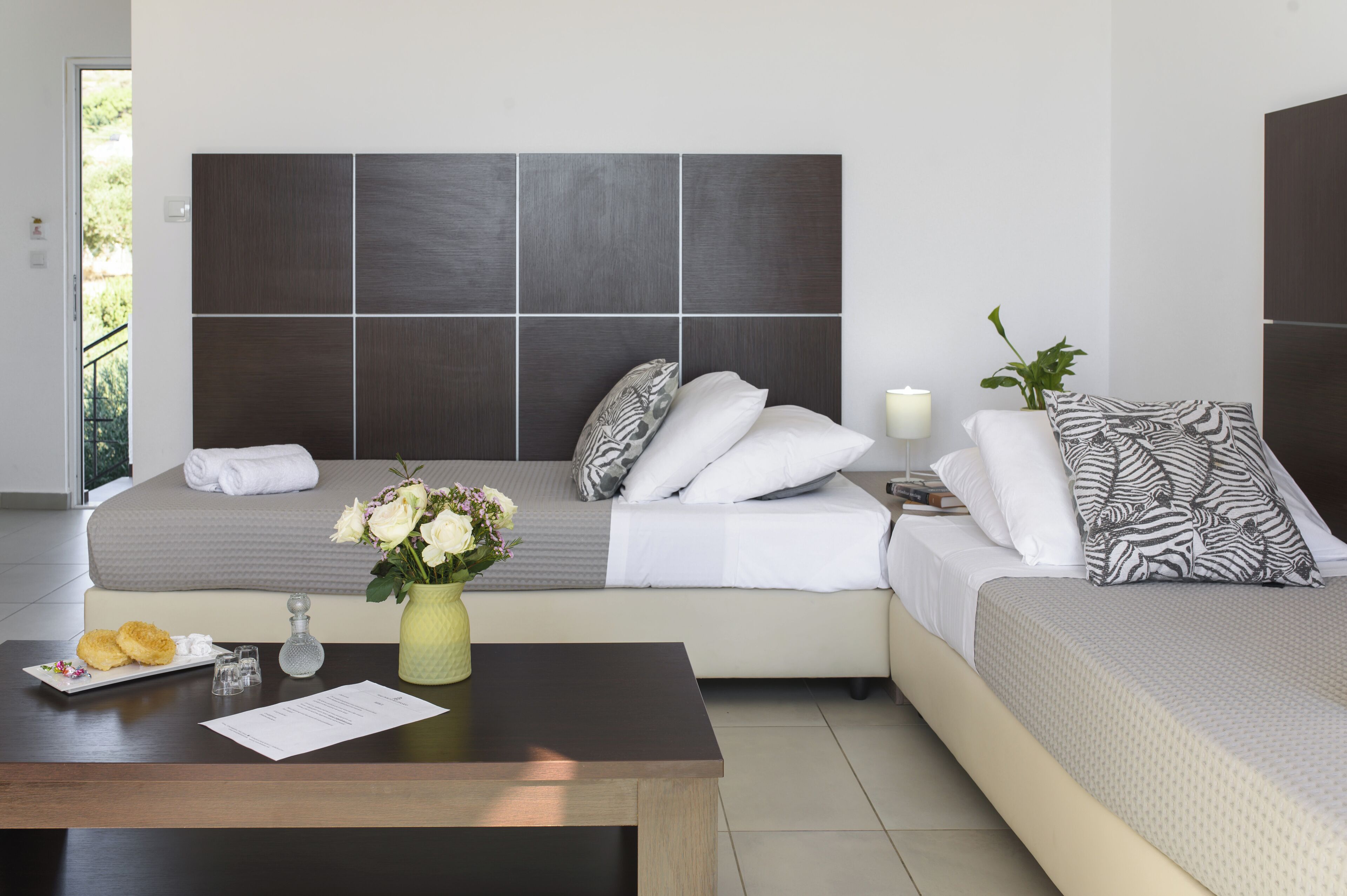 Una habitación de hotel moderna con una cama grande, una mesita de noche con un jarrón de flores y una pequeña mesa con una bandeja de pasteles.