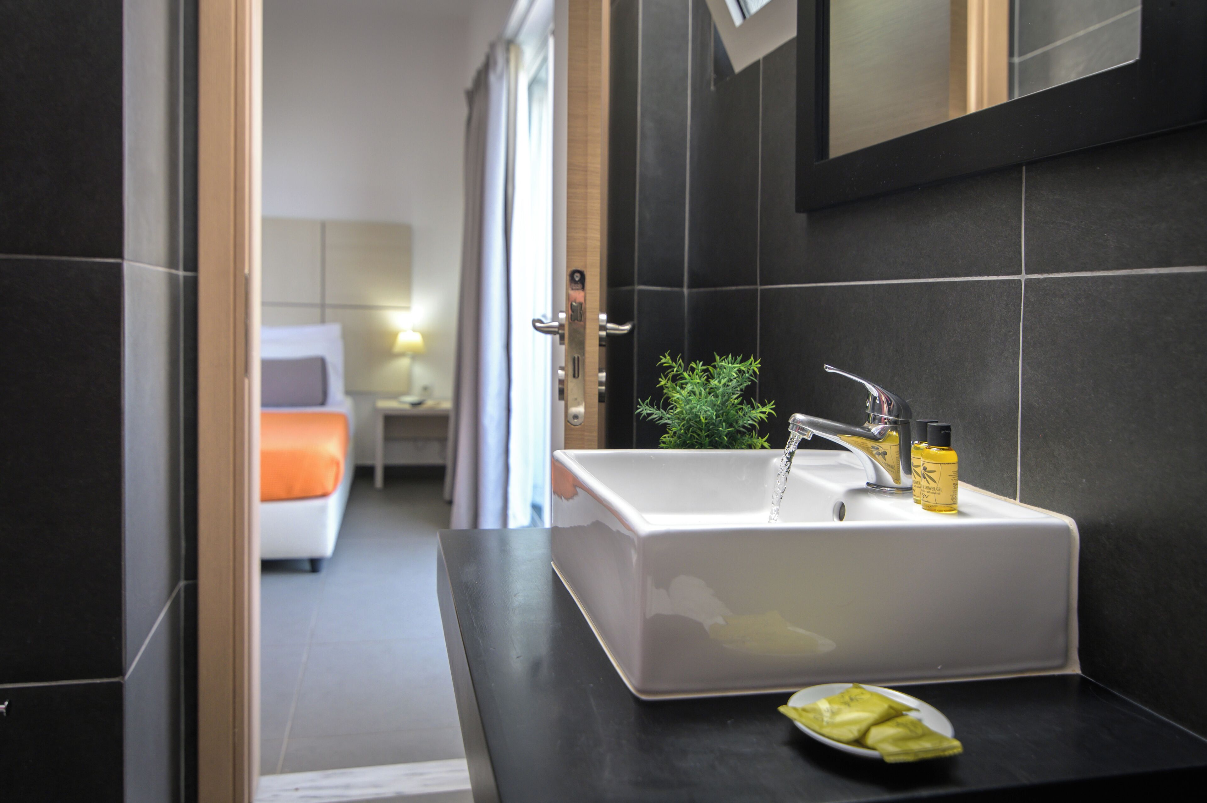 A modern bathroom with a white sink, dark tiled walls, a small potted plant, and a bottle of lotion on the counter.