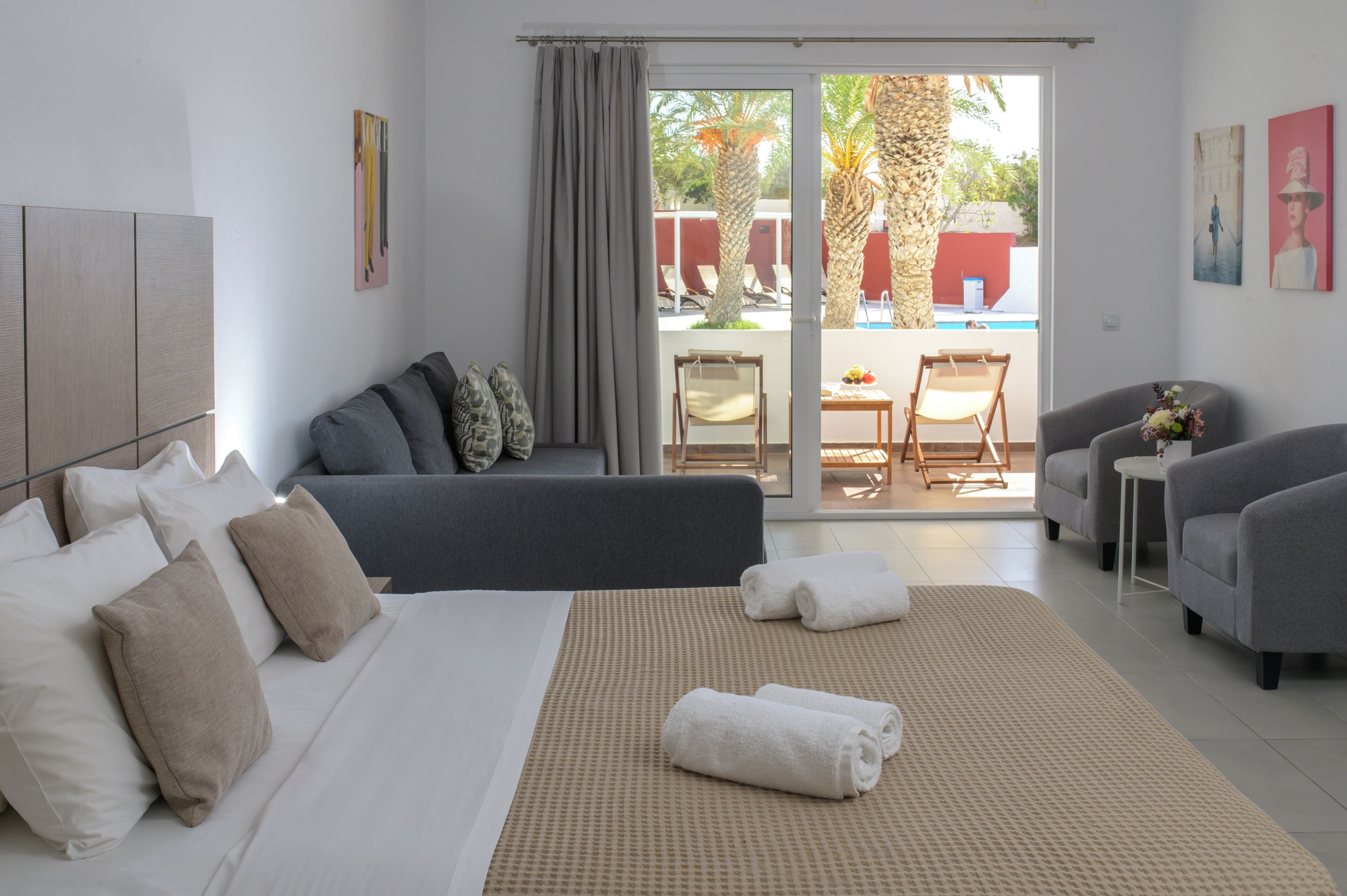 A modern hotel room with a bed, grey sofa, and a balcony with a pool view.