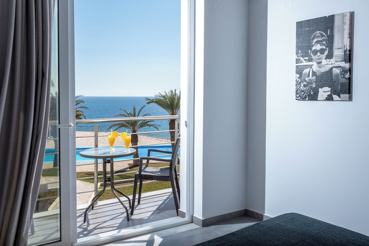 Un balcon avec vue sur la mer, une table en verre, deux chaises et un tableau encadré au mur.