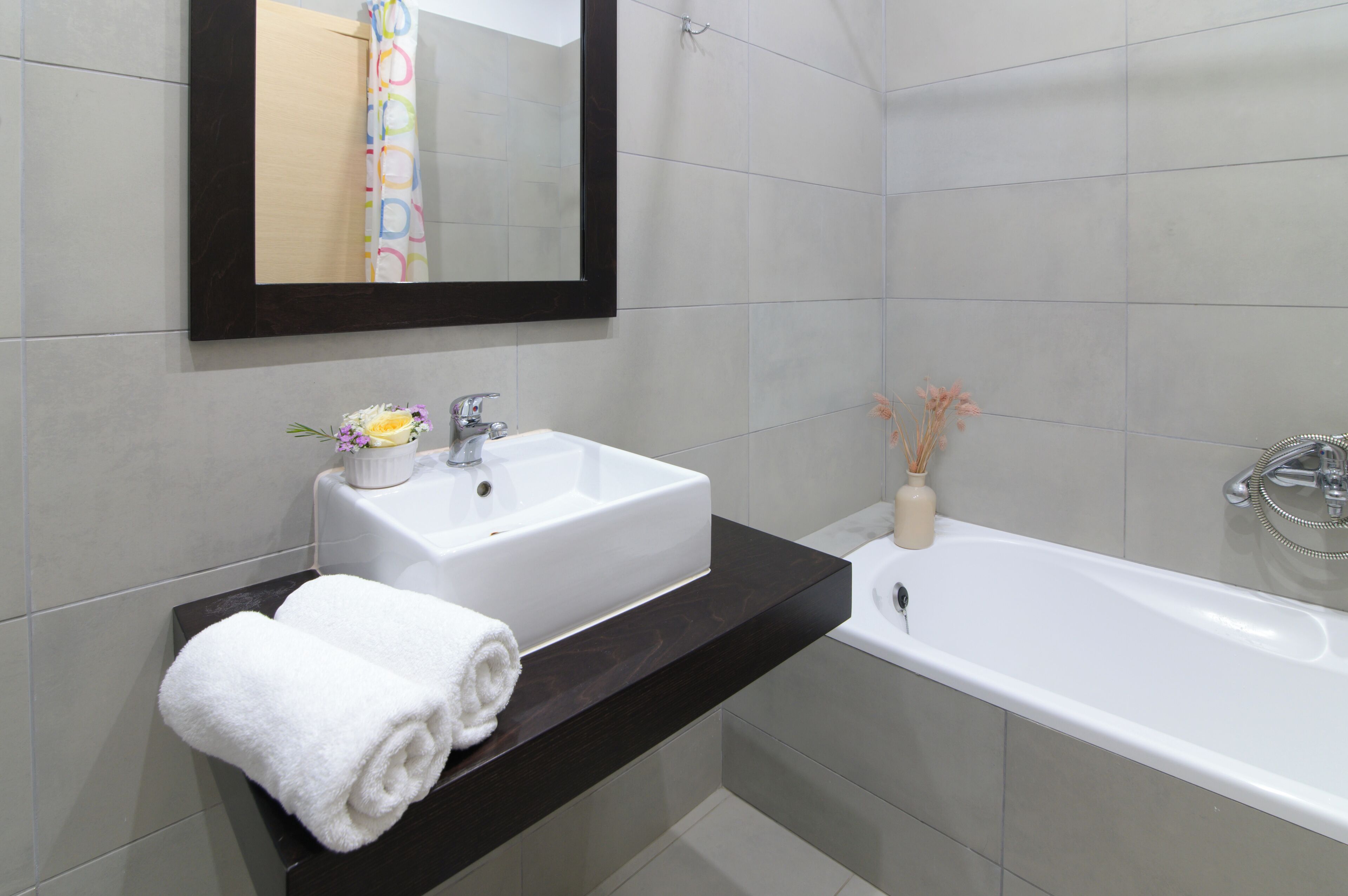Un baño con un lavamanos blanco, un espejo y una bañera. Hay toallas en la encimera y un florero con flores.