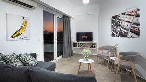 Un salon moderne avec un canapé gris, un téléviseur à écran plat, une bibliothèque et une grande fenêtre donnant sur le coucher de soleil.
