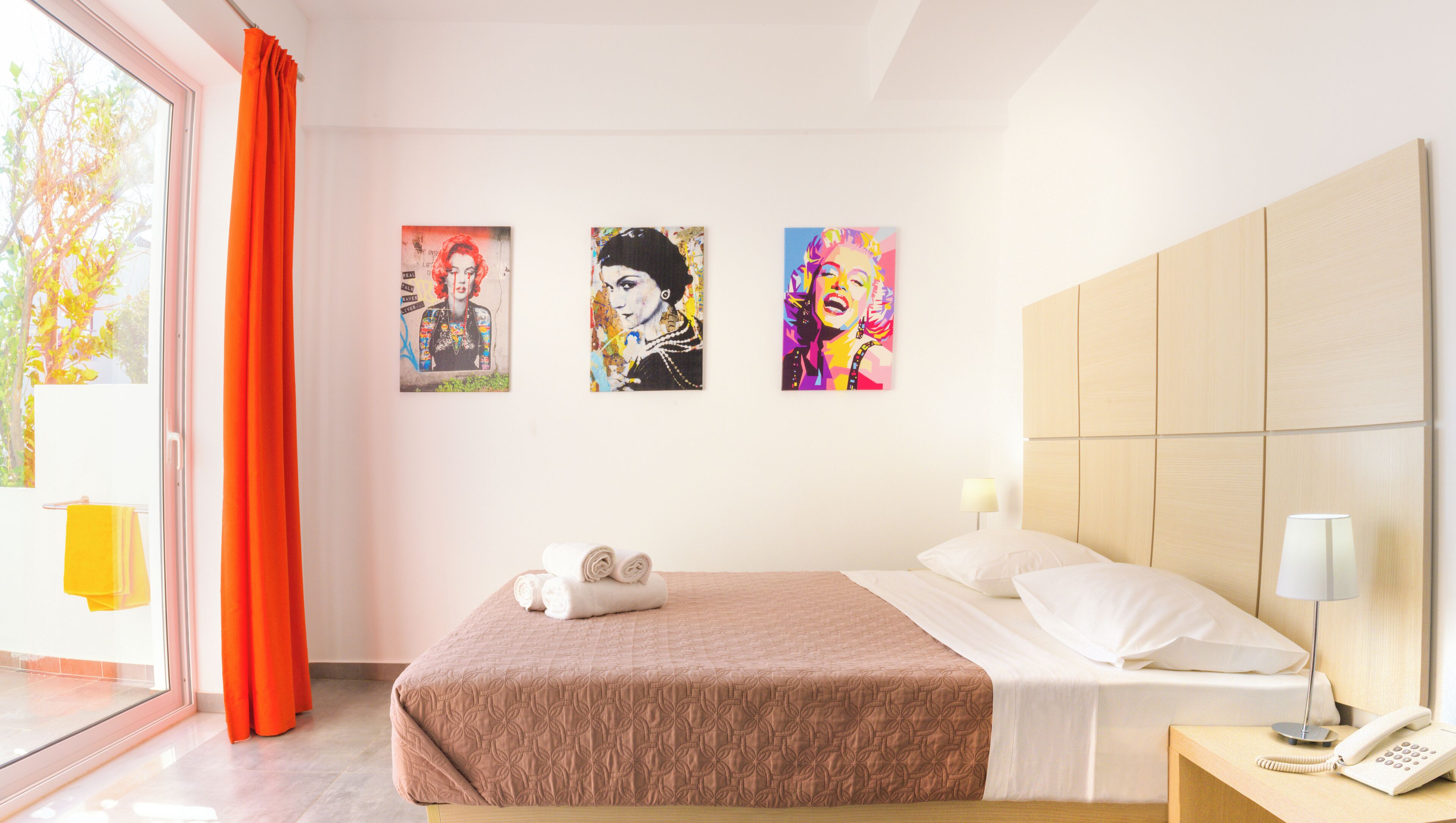 A hotel room with a bed, a nightstand, a telephone, and three artworks on the wall.
