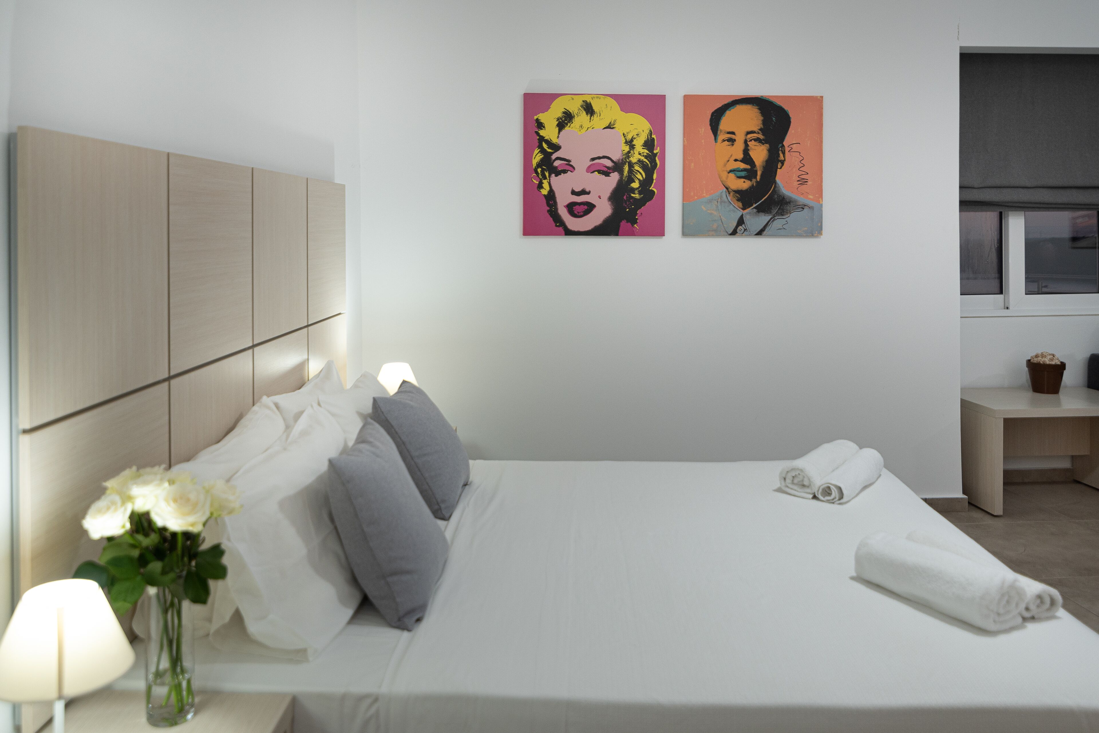 A neatly made bed with pillows, a vase of flowers, and two paintings on the wall.