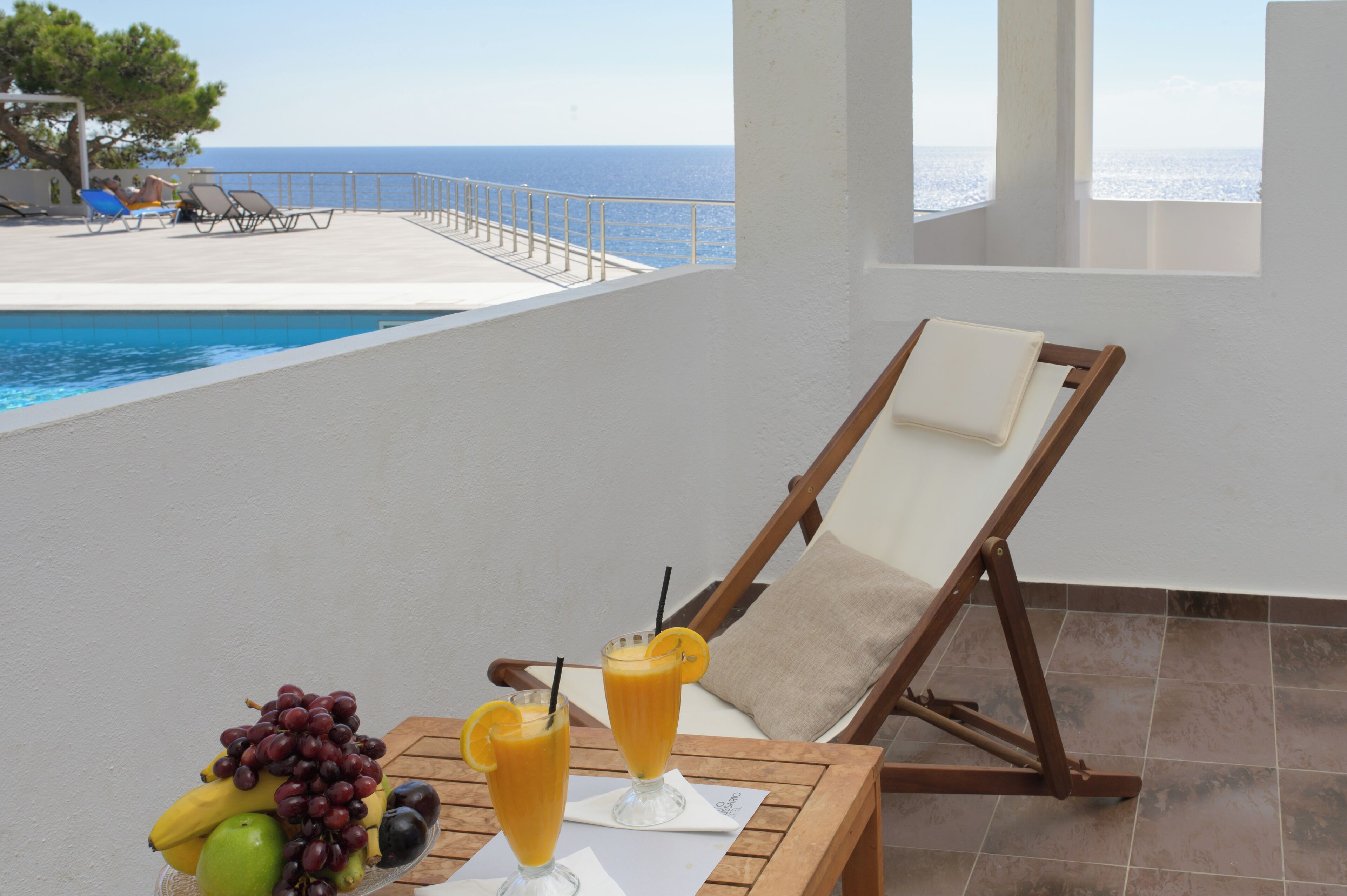 A poolside area with a lounge chair, a table with fruit and drinks, and a view of the sea.