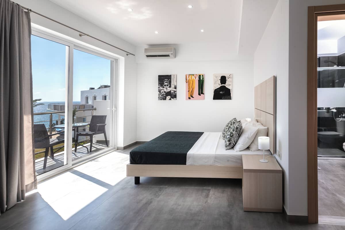 Une chambre moderne avec un grand lit, une table de chevet et une vue sur la mer grâce à de grandes fenêtres du sol au plafond.