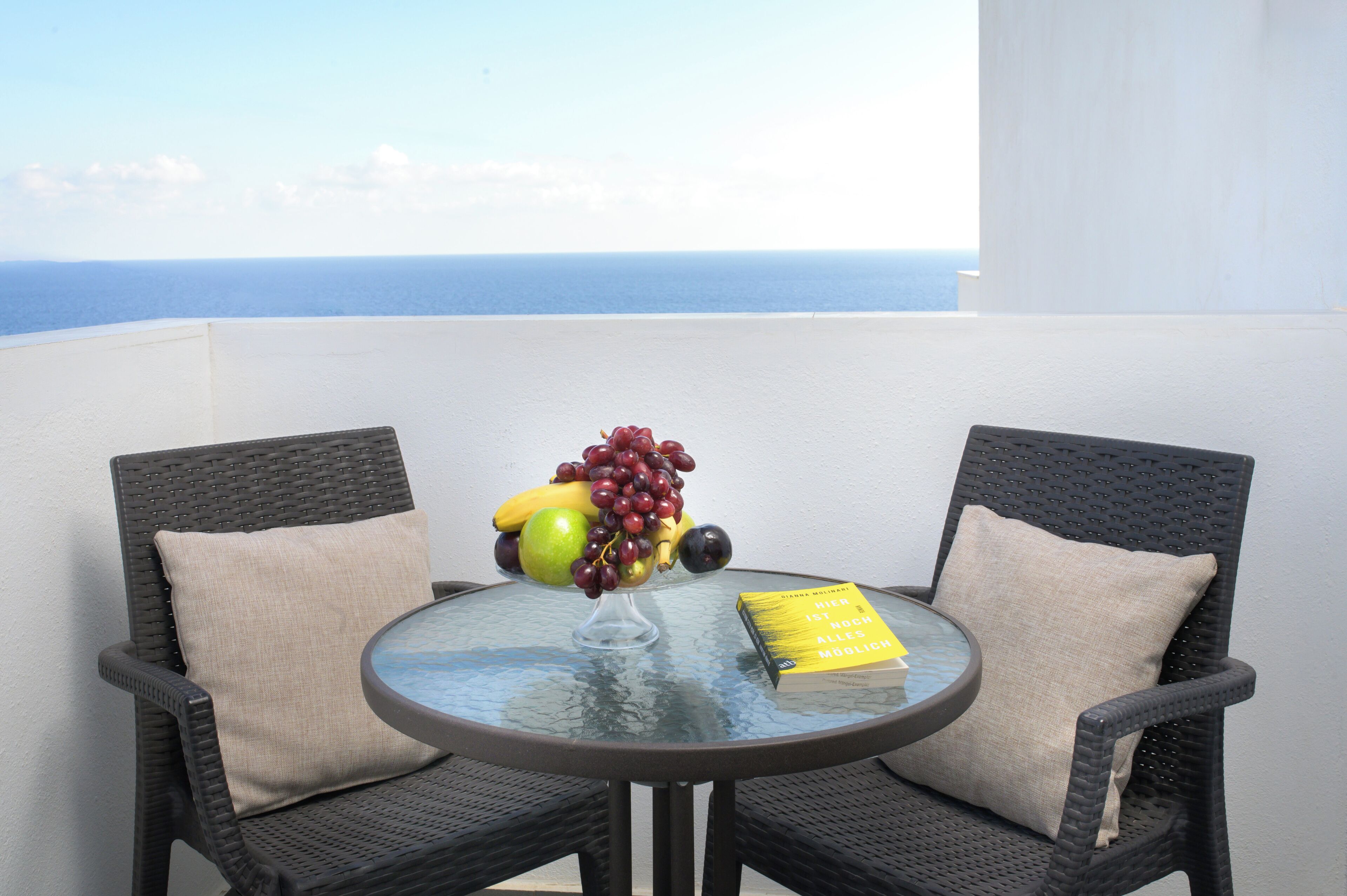 A round glass table with a fruit bowl and a book, surrounded by wicker chairs with cushions, overlooking a view of the sea.