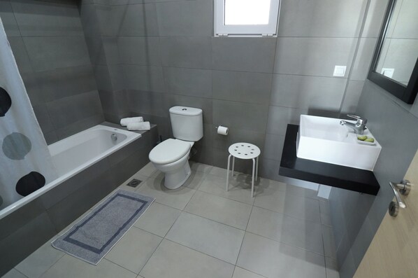 A modern bathroom with a bathtub, toilet, sink, and a small stool. - Porto Belissario (Ierapetra)