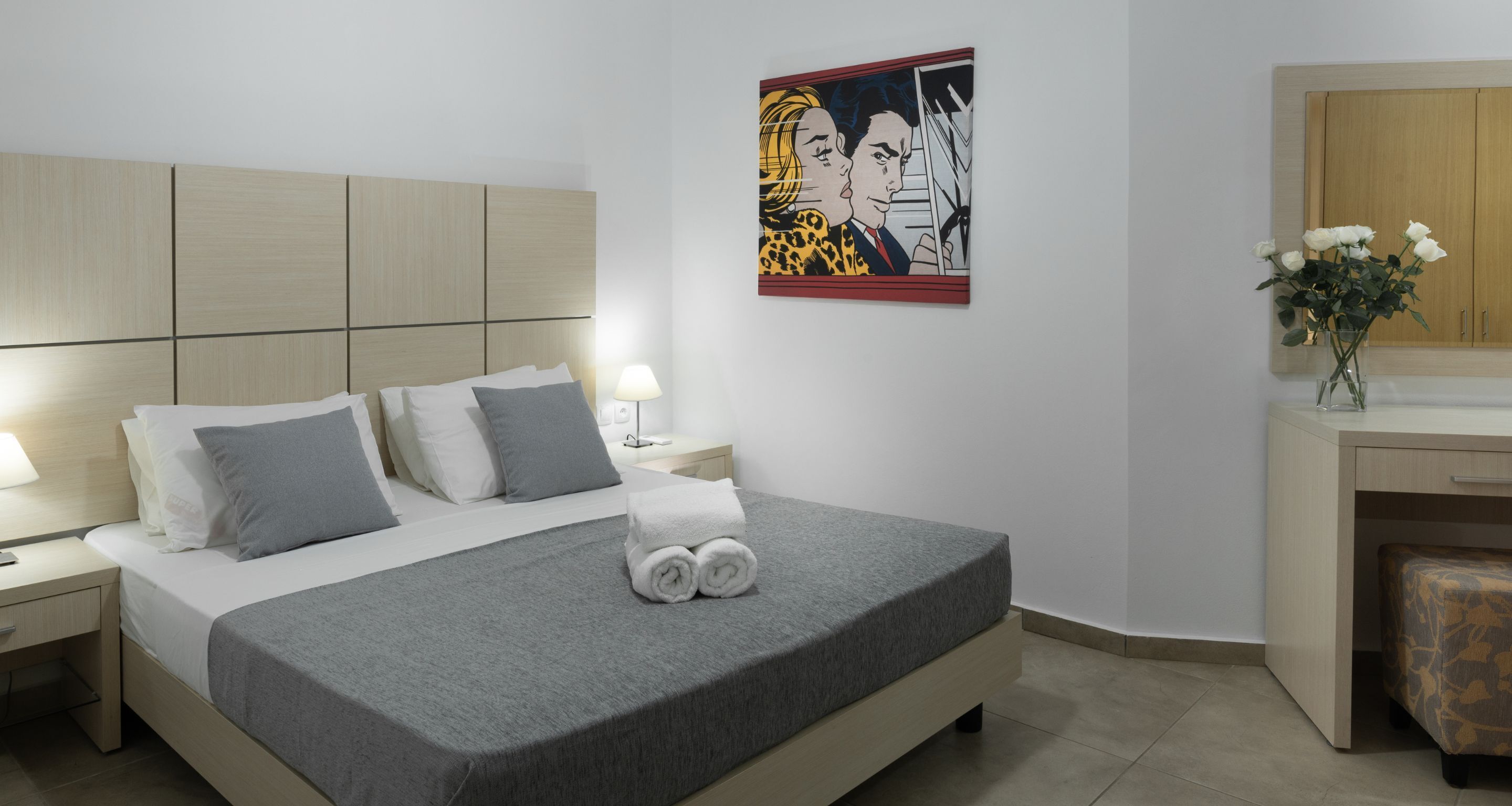 Una habitación de hotel moderna con una cama grande, una mesita de noche con una lámpara, un cuadro en la pared y un escritorio con un florero.