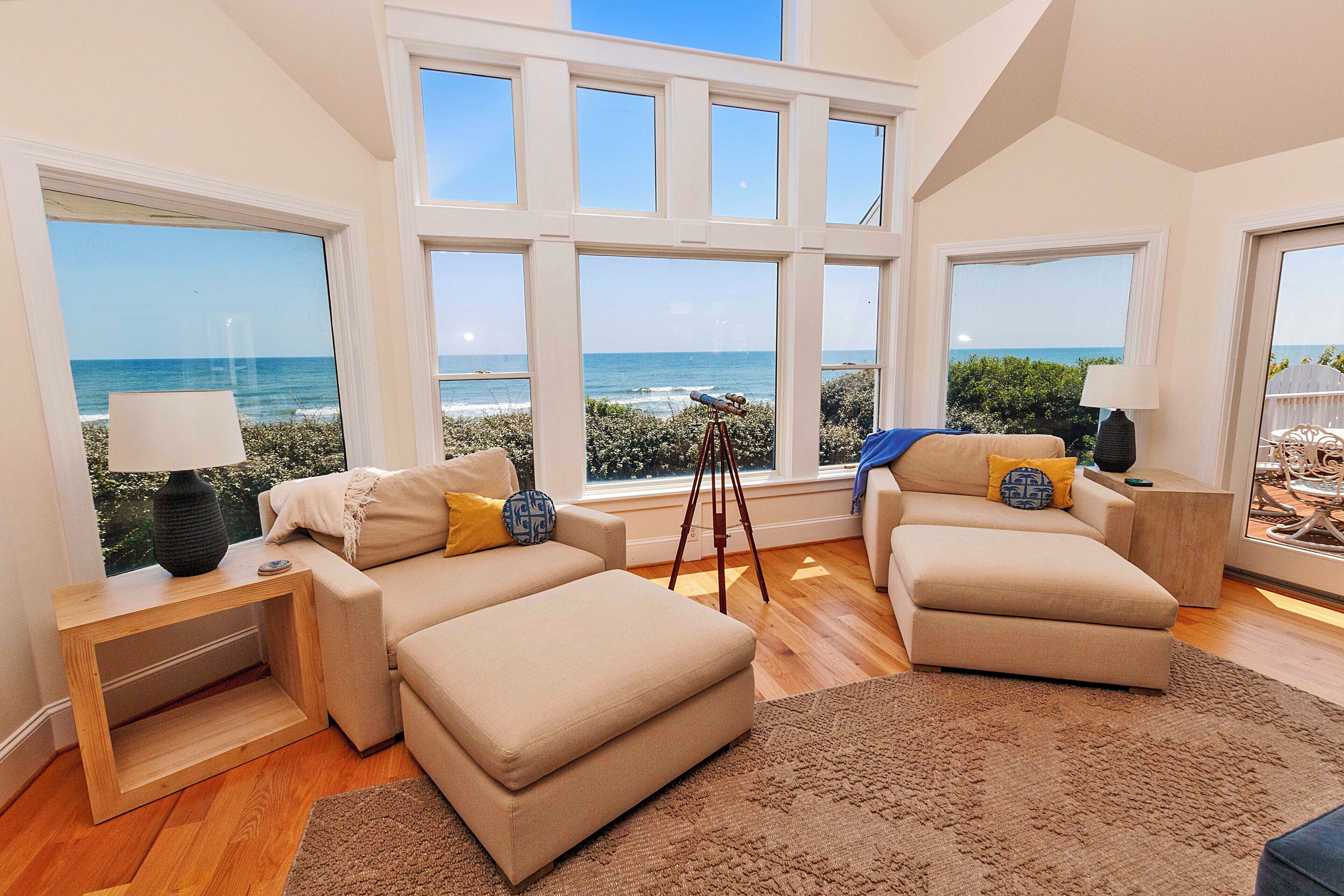 A spacious living room with large windows offering ocean views, a cozy sofa, a wooden side table, and a tripod with a telescope.