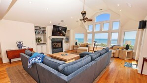 A spacious living room with a large gray sectional sofa, a wooden coffee table, and a fireplace. There is a television mounted above the fireplace and a bookshelf filled with books. The room has large windows offering a view of the ocean. - OCEANFRONT, Ocean views, ELEV, Private Pool, Private beach access, WL1083, Lighthouse Drive (Corolla)