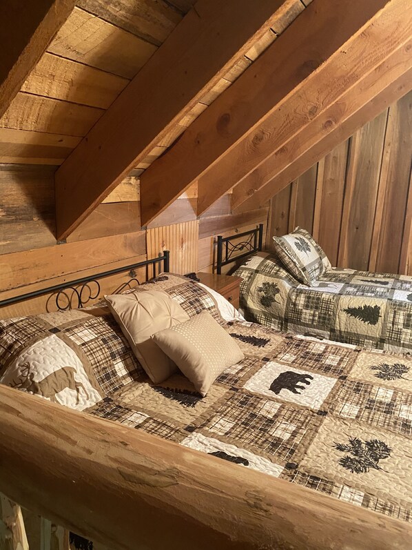 A loft-style bedroom with a sloped ceiling, wooden beams, and a bed with a quilted bedspread and multiple pillows.