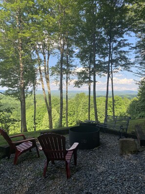 A seating area with two Adirondack chairs and a fire pit, surrounded by trees and a view of a forested landscape.