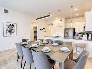 A modern kitchen with a central island, stainless steel appliances, and a dining area with grey chairs and a table set for four. - Reunion Resort Condo: Waterpark, Clubhouses and fun! (Four Corners)