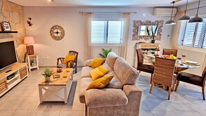 A modern living room with a flat-screen TV, sofa, dining table, and a decorative wall clock. - Casa Balimar - Pool, Jacuzzi, Internet, Garten, Meerblick (Tarajalejo)