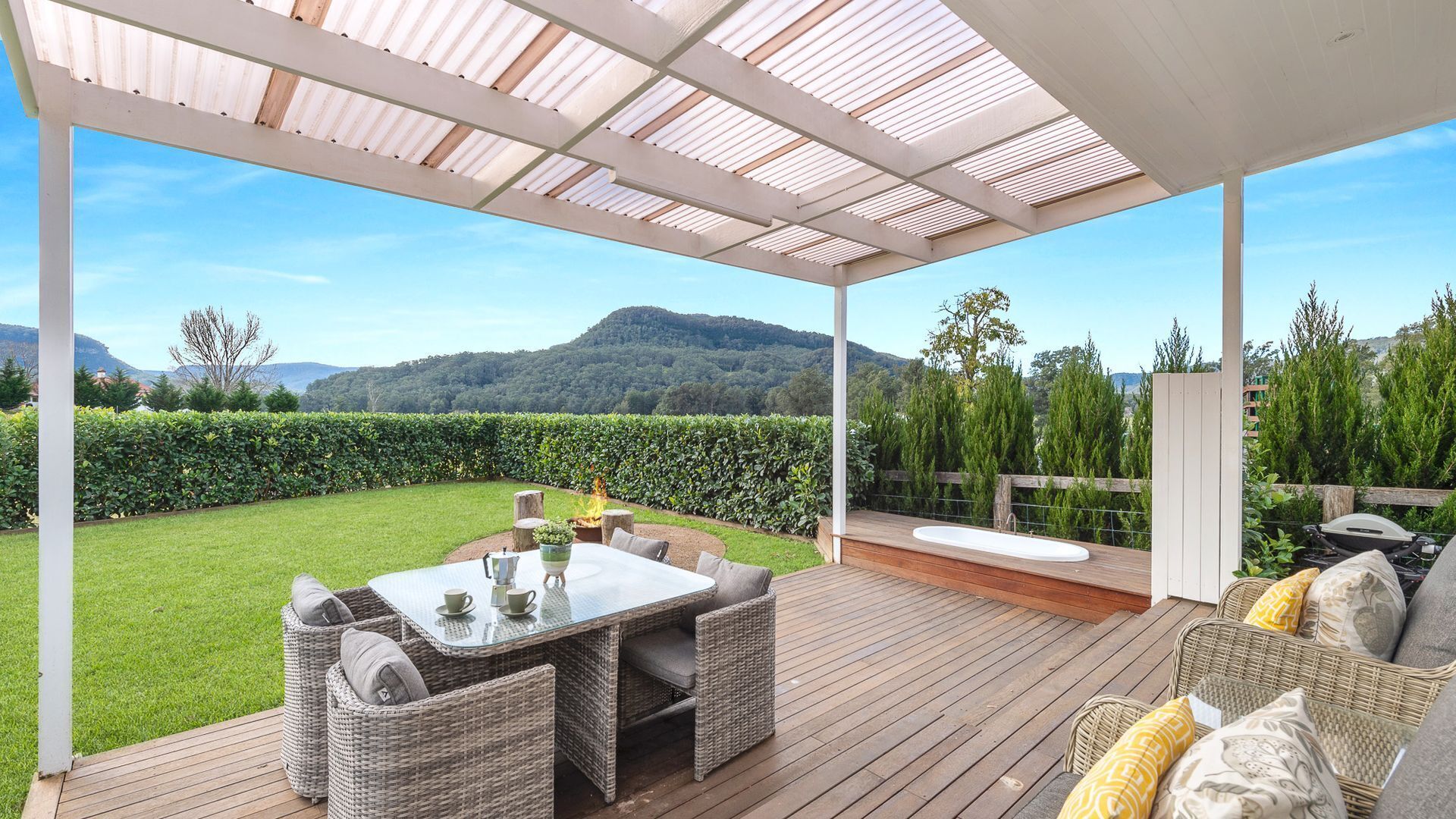 A covered outdoor patio with a table and chairs, a hot tub, and a view of a lush garden and mountains.