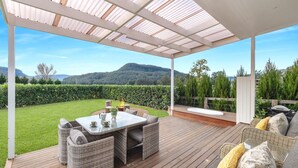 Un patio techado al aire libre con una mesa y sillas, un jacuzzi y vistas a un jardín exuberante y montañas.