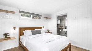 Un dormitorio moderno con cama de madera, ropa de cama blanca, una mesita de noche con una planta y vistas al baño a través de una puerta abierta.