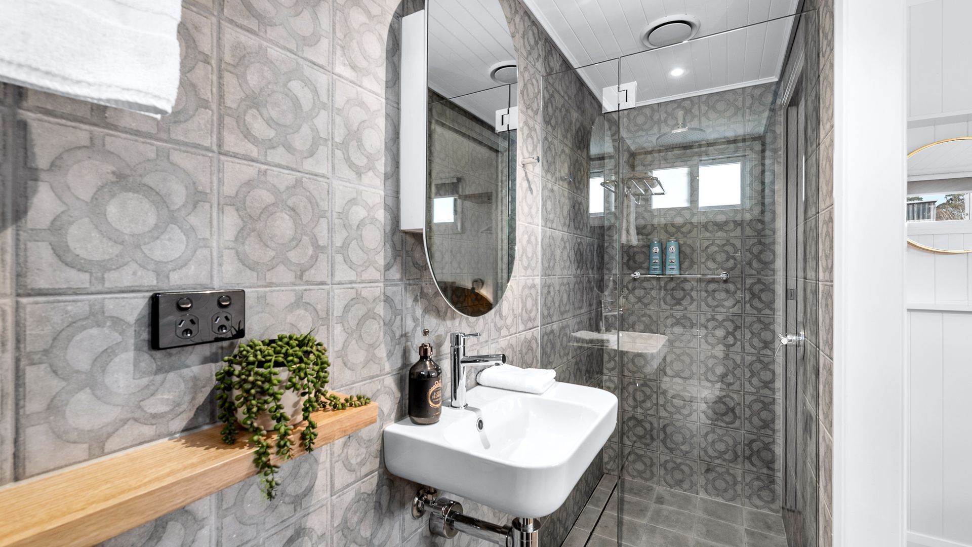 A modern bathroom with patterned tiles, a white sink, a mirror, a shower enclosure, and a plant on a shelf.