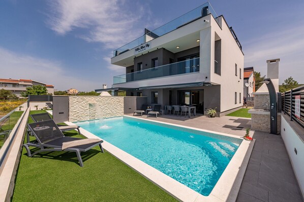 A modern two-story house with a swimming pool, outdoor seating, and a stone wall.