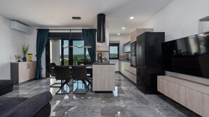 Una cocina moderna con un televisor grande, una zona de comedor con mesa y sillas, y vistas al exterior a través de amplias ventanas.