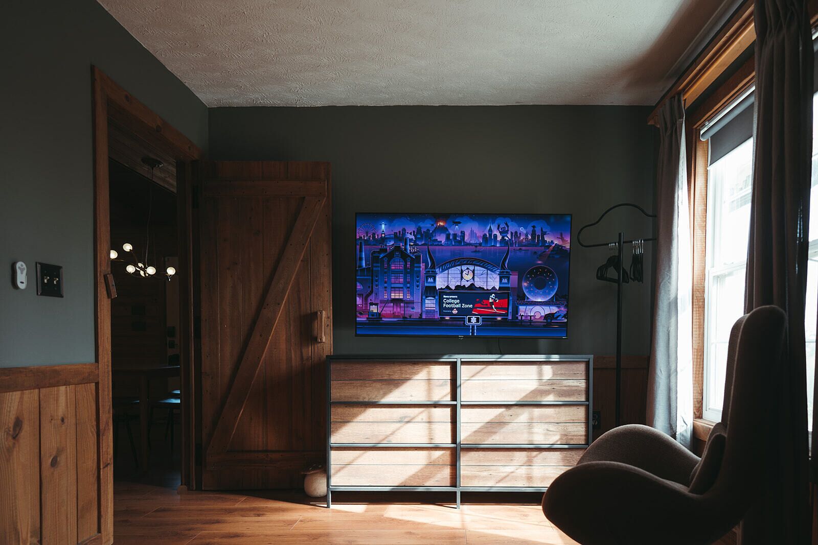 Ein Zimmer mit einem großen Flachbildfernseher an der Wand, Holzboden, einer Schiebeglastür und einem gepolsterten Sessel.