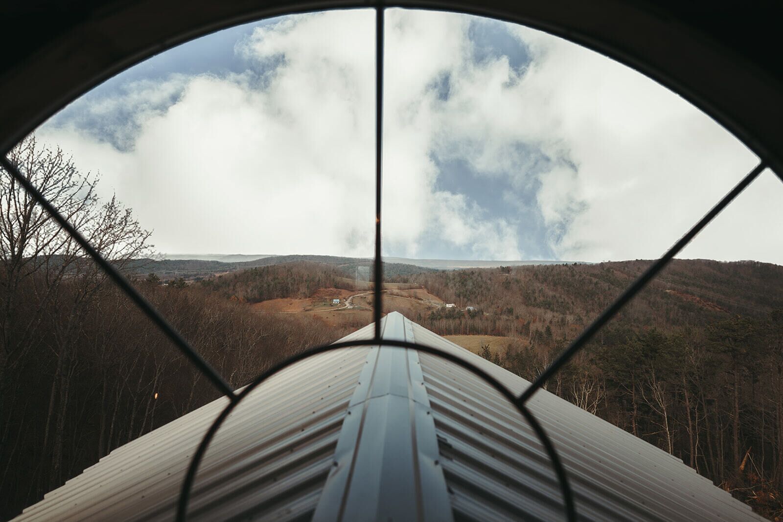 Une vue à travers une fenêtre à cadre métallique, donnant sur une vallée parsemée d’arbres et où l’on aperçoit un bâtiment au loin.