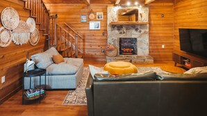 A cozy log cabin interior with a stone fireplace, a sofa, a television, and a staircase. - Endless Views: Sunsets, Sauna, Outdoor Firepit under the Stars (Max Meadows)