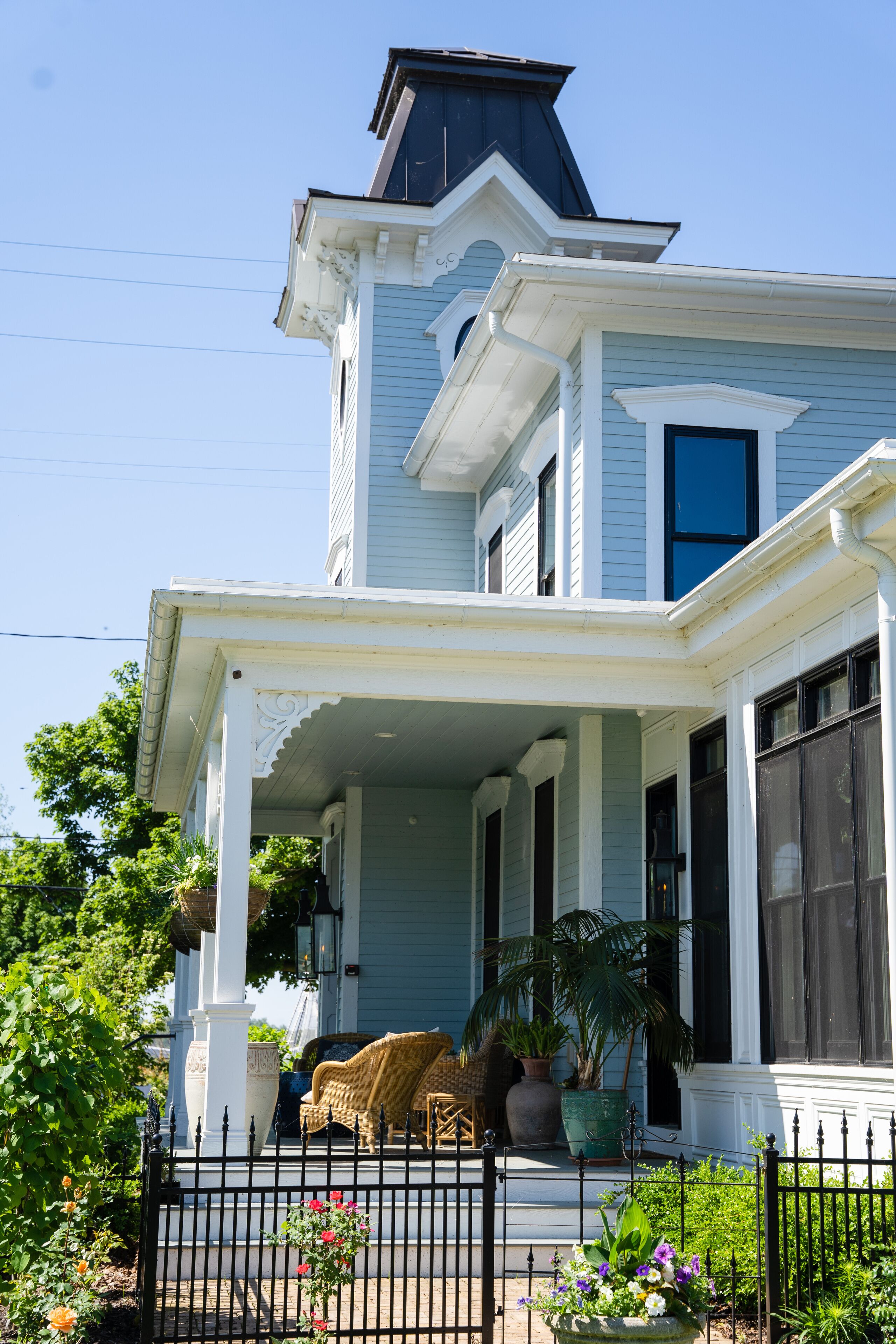 A light blue Victorian-style house with a porch, potted plants, and a black metal fence.