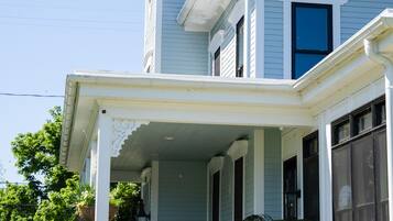 A light blue Victorian-style house with a porch, potted plants, and a black metal fence.