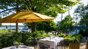 An outdoor dining area with tables and chairs under a large umbrella, surrounded by greenery and a view of the water. - The Lilley Mansion Bed & Breakfast (Spring Lake)