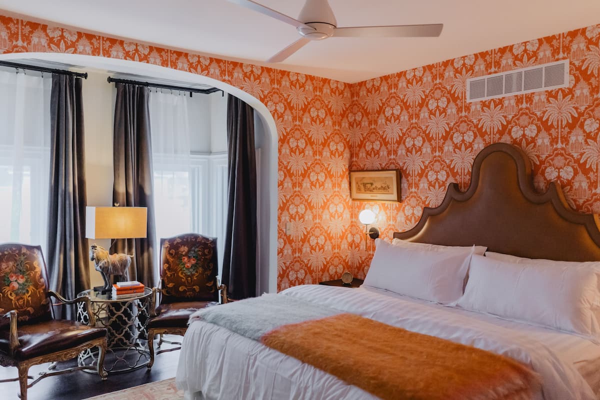 Hotellihuone, jossa on suuri sänky, kaksi nojatuolia, pieni pöytä lampun kanssa ja kattotuuletin.