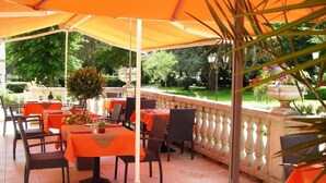 Un'area pranzo all'aperto con tavoli e sedie sotto un'ampia tenda arancione, circondata da vegetazione e un giardino curato.