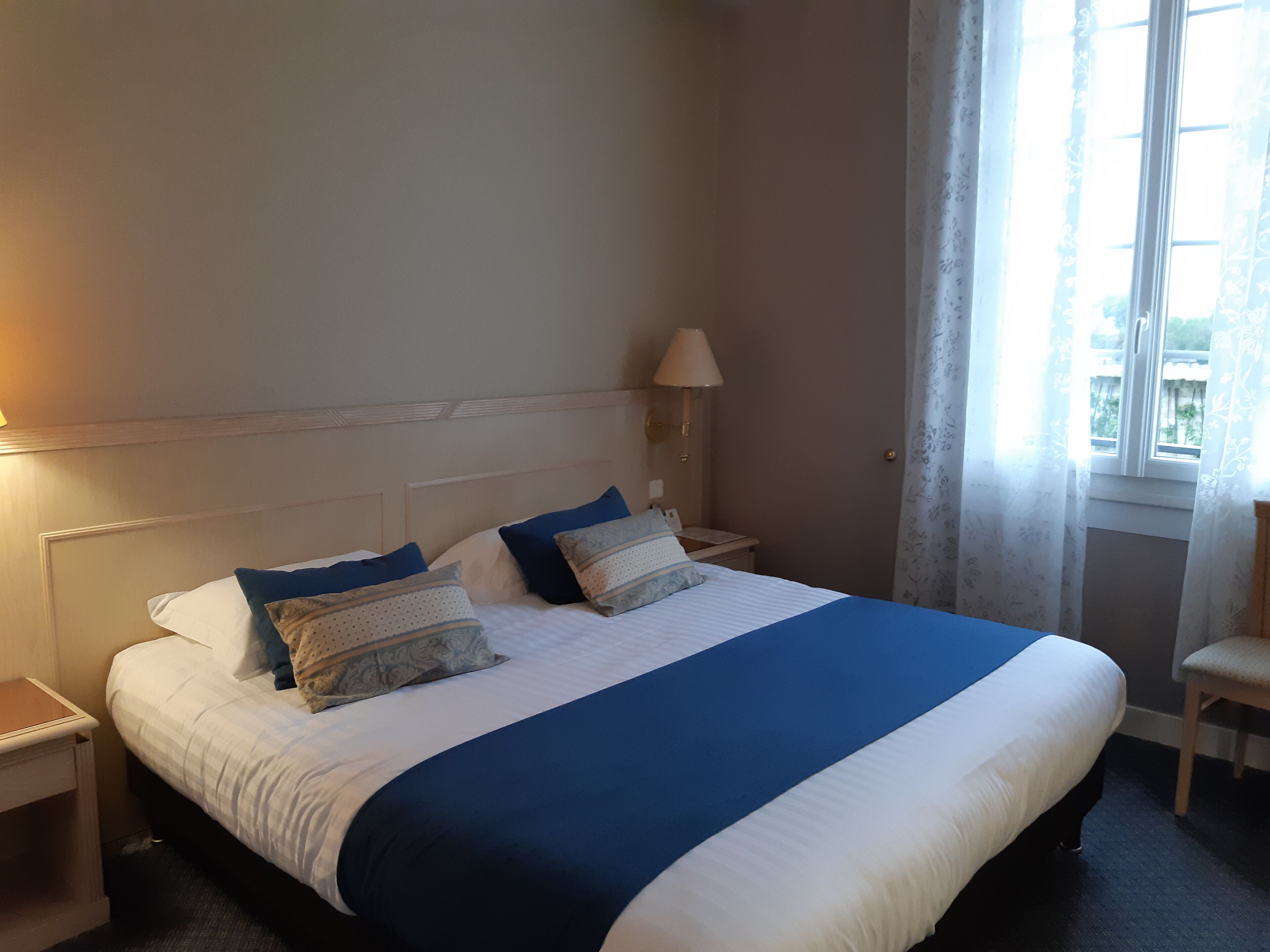Una cama bien hecha con una colcha azul y blanca, dos mesitas de noche con lámparas y una ventana con cortinas.