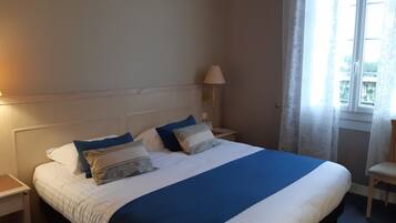 Una cama bien hecha con una colcha azul y blanca, dos mesitas de noche con lámparas y una ventana con cortinas.