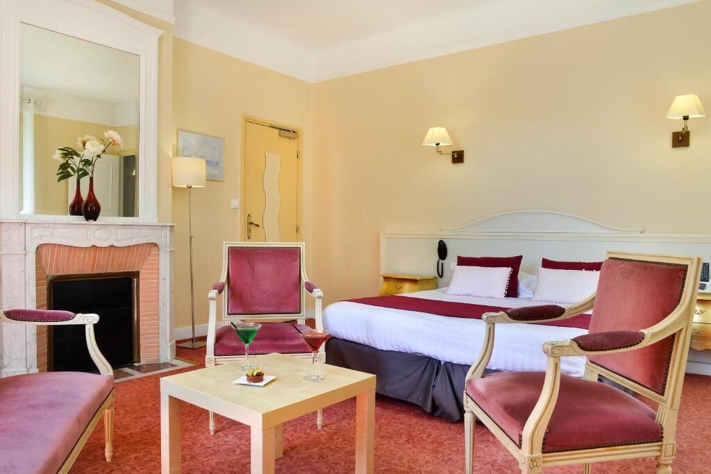 A hotel room with a bed, two armchairs, a small table, a fireplace, and a mirror.