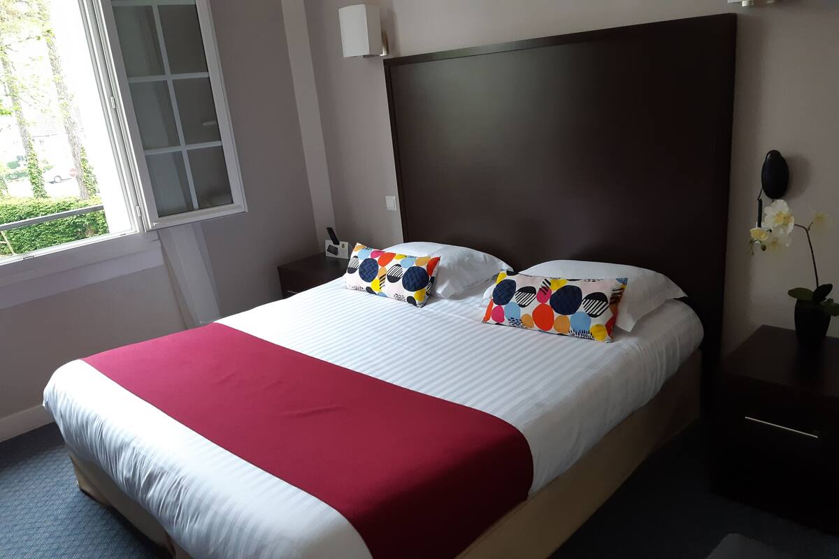 Una cama bien hecha con colcha roja y blanca, dos almohadas con diseños coloridos, un cabecero oscuro, una mesita de noche con una lámpara y una ventana con vistas a la vegetación.