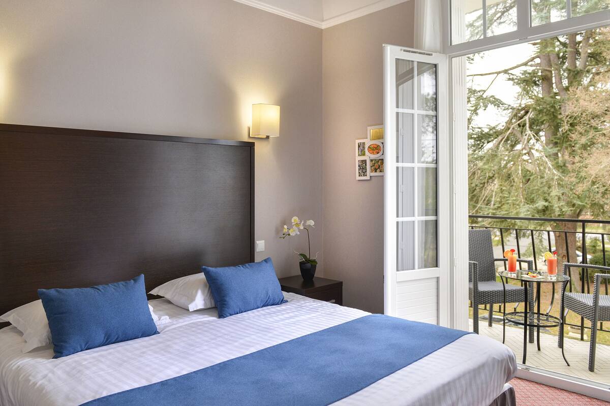 Habitación de hotel con una cama grande, una mesita de noche con lámpara, una orquídea en maceta y un balcón con mobiliario exterior.