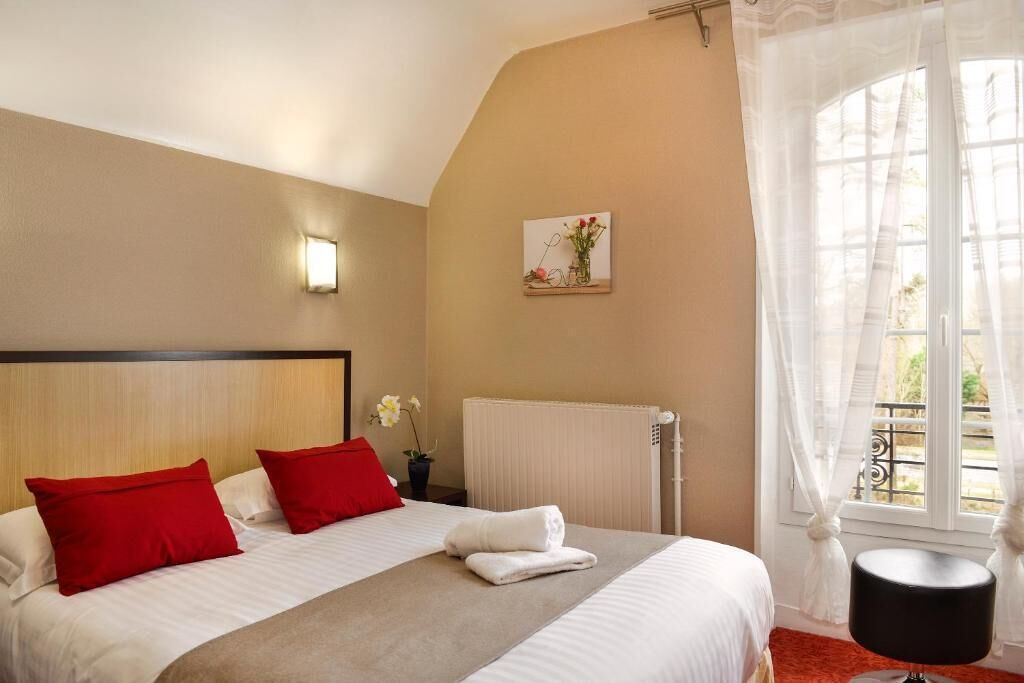Habitación de hotel con una cama grande, almohadas rojas y blancas, una mesita con un florero, un radiador y una ventana con cortinas traslúcidas.