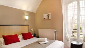 Habitación de hotel con una cama grande, almohadas rojas y blancas, una mesita con un florero, un radiador y una ventana con cortinas traslúcidas.
