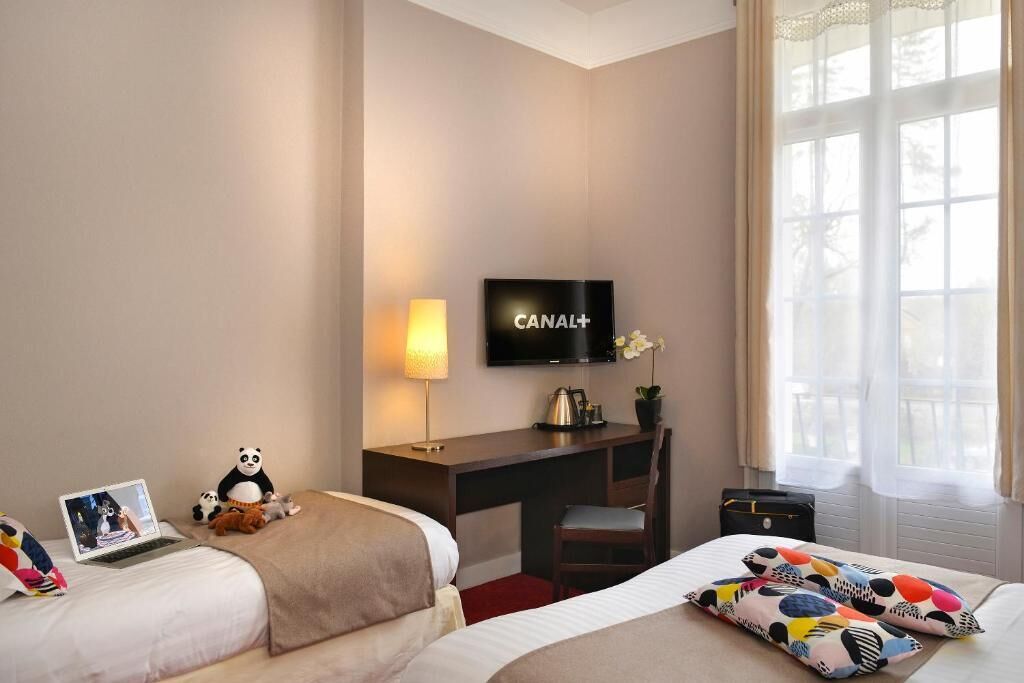 A hotel room with two beds, a desk with a TV, a lamp, and a window with curtains.