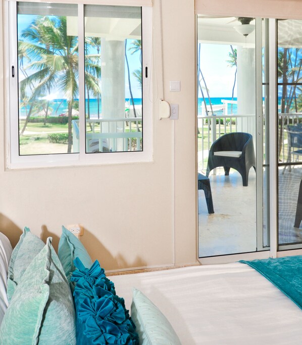 Ein Zimmer mit einem Bett, einem Fenster mit Blick auf Palmen und das Meer, sowie einer Glastür, die zu einem Außenbereich mit Stühlen führt.