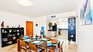 Un coin repas avec une table en bois dressée pour quatre personnes, entourée de chaises. À gauche, on aperçoit une bibliothèque noire remplie de livres et d’objets décoratifs. On distingue également, au fond, un espace cuisine équipé d’appareils modernes et d’un distributeur d’eau.