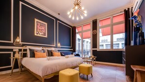 A modern hotel room with a large bed, two yellow ottomans, a chandelier, and a view of the city through large windows. - The Blue Elysée (Paris)