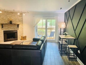 En moderne stue med pejs, en sort lædersofa, et spiseområde med bord og stole, og udsigt til det fri gennem en glasdør.