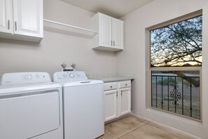 A laundry room with two washing machines, a dryer, and a window with a view of trees and a fence. - Tierra Velo - HGTV Style in East Tucson (Tucson)