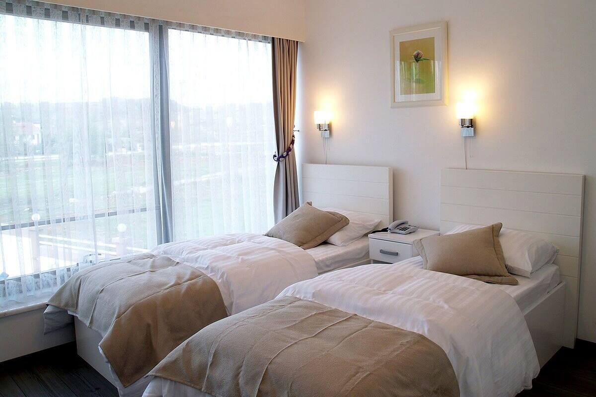 A hotel room with two beds, a window with curtains, a bedside table with a phone, and a wall-mounted light fixture.